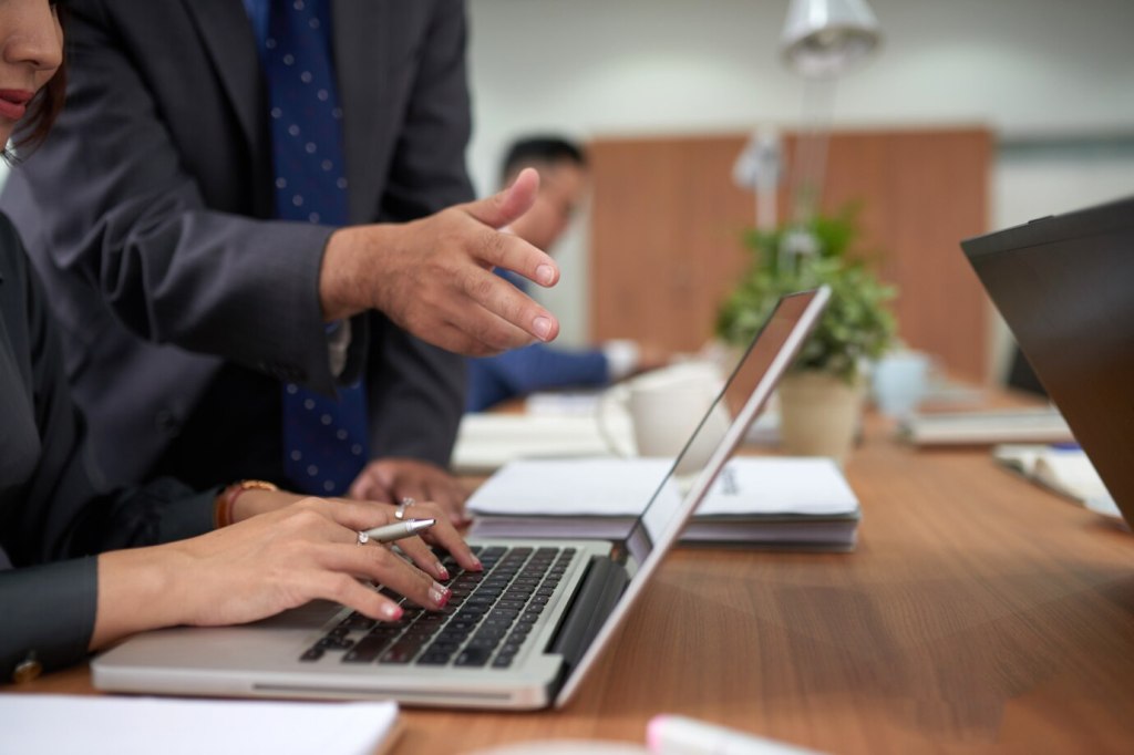 unrecognizable-woman-working-laptop-office-man-standing-watching-pointing
